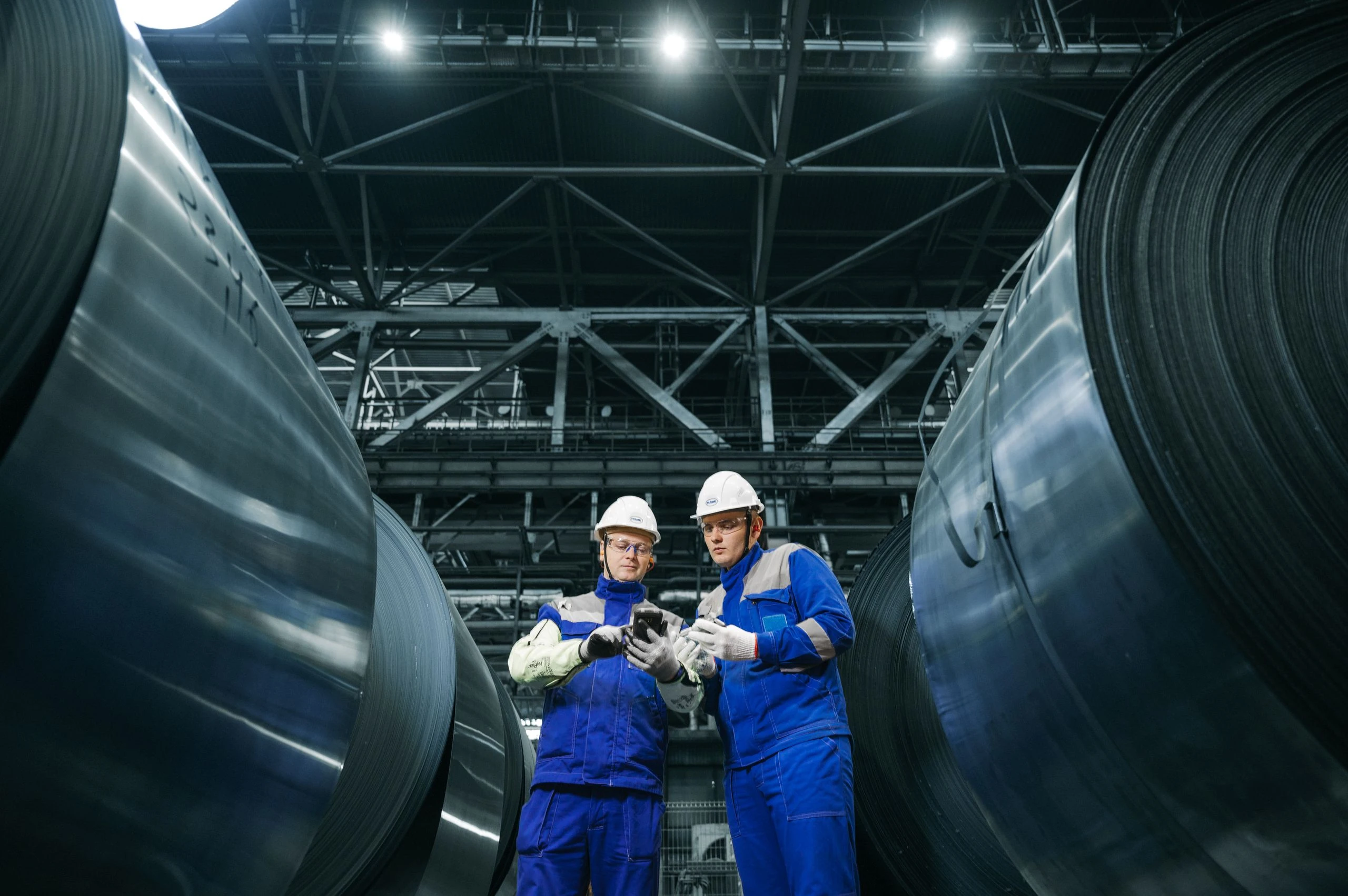 Two industrial specialists reviewing a tablet beside large steel coils